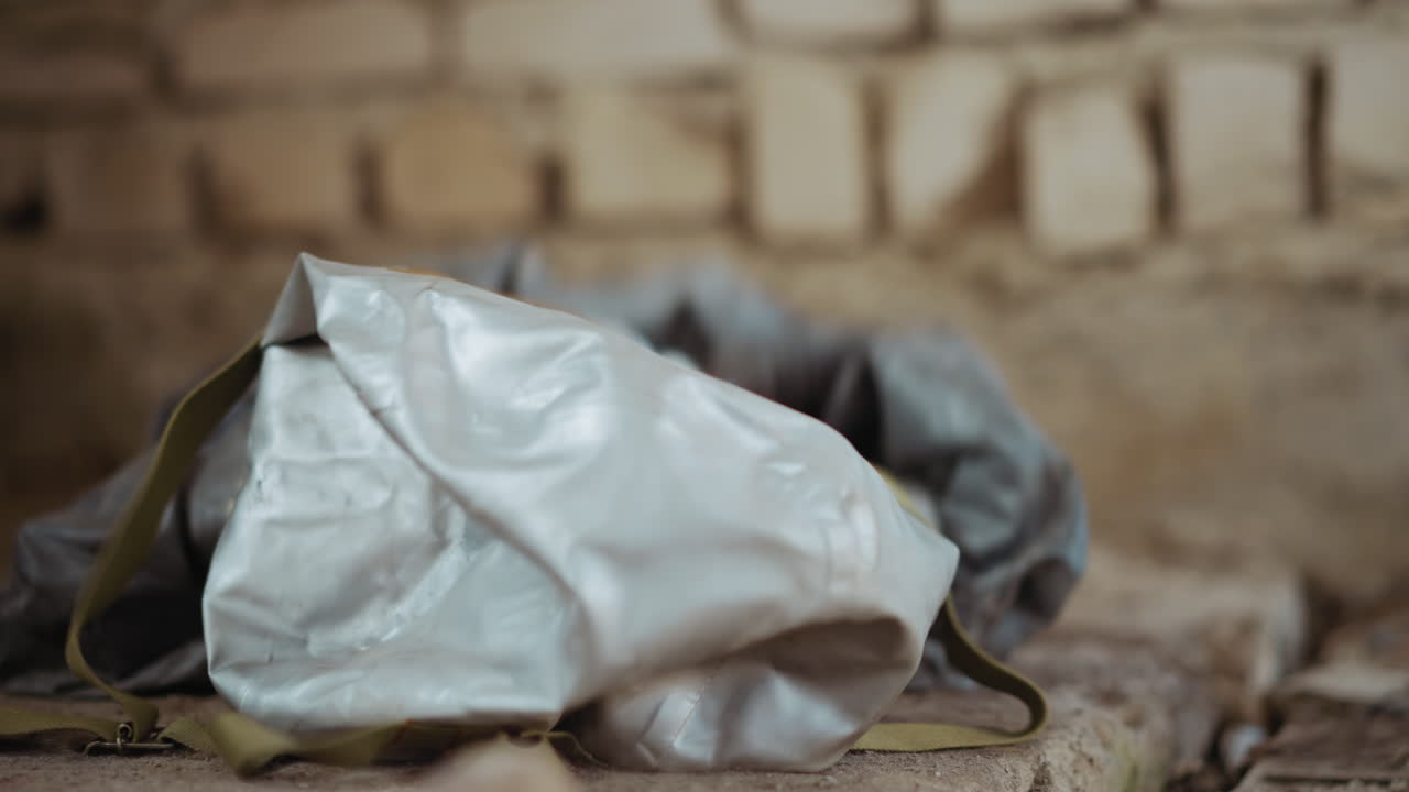 Close up of person dropping protective hazmat mask onto rough dusty surface with cracked concrete, faded background bricks, muted tones and somber mood suggesting danger