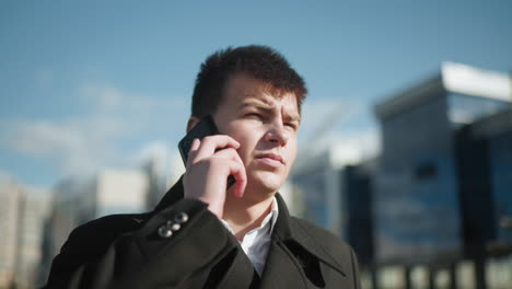 investor in black coat answering phone call outdoors in urban setting, background features blurred modern buildings and greenery under bright sunlight, exuding professionalism