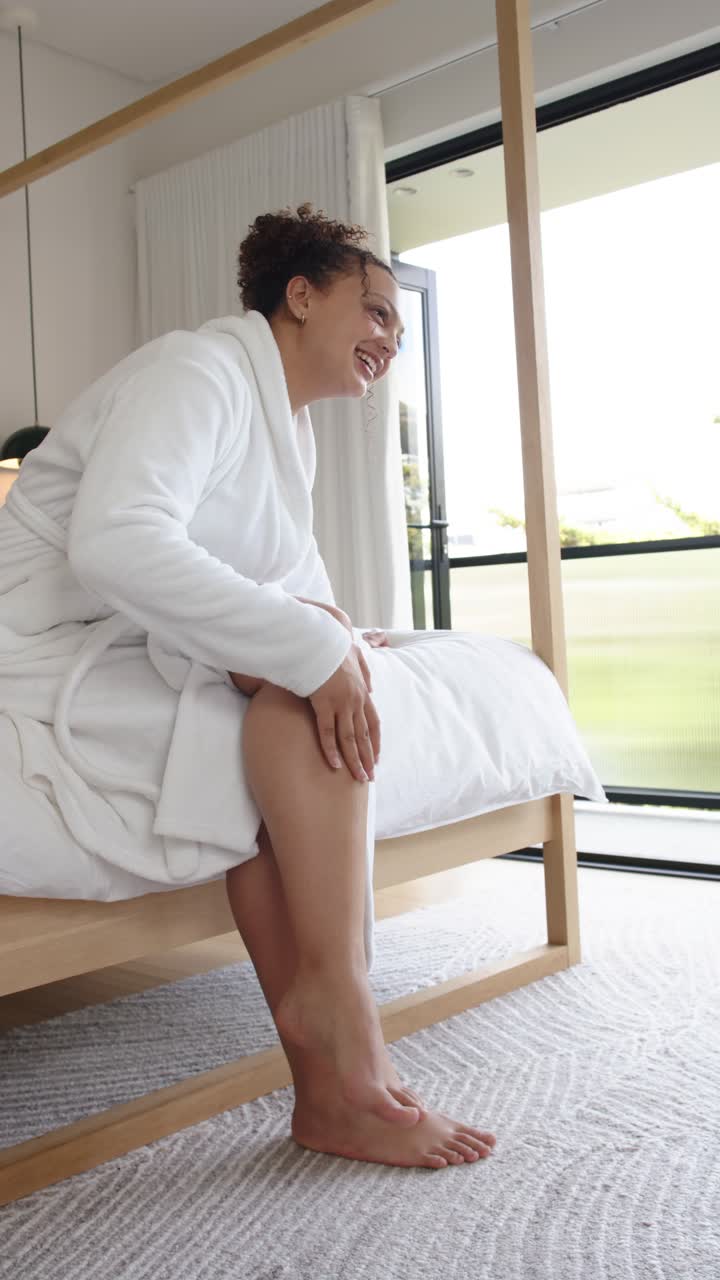 Vertical video: African American woman in bathrobe sitting on bed smiling at sunlight rubbing ankle