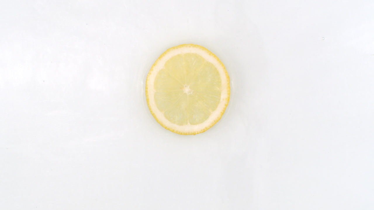 On a white background a splash of water falls on a slice of lemon in slow motion.