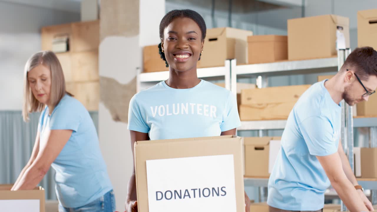 voluntaria afroamericana sosteniendo cajas de donación y mirando la cámara en un almacén de caridad mientras sus compañeros de trabajo trabajan en cajas de embalaje