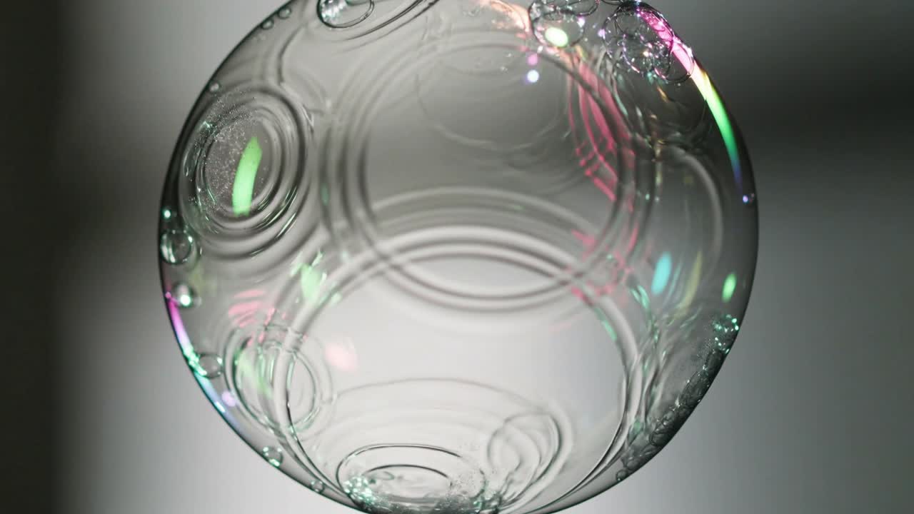 Bolla di sapone che fluttua e affonda su sfondo grigio per via di una brezza, mostrando increspature, goccioline, verde e rosa.