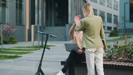 portrait of attractive couple of man and woman sitting on bench by modern office building in smart casual business style, working together on laptop, smiling. outside, freelance worker concept