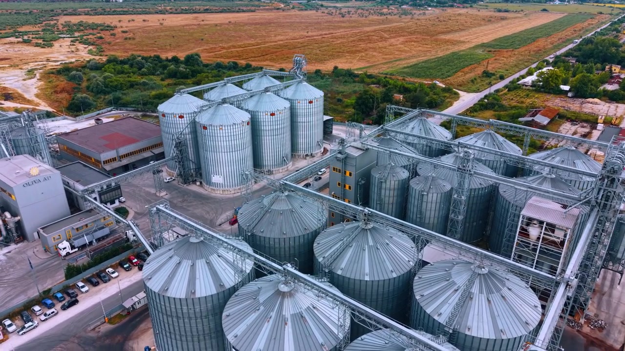 Agricultural grain elevator with metallic storage silos. Steel bins stand near rural fields in bright daylight.