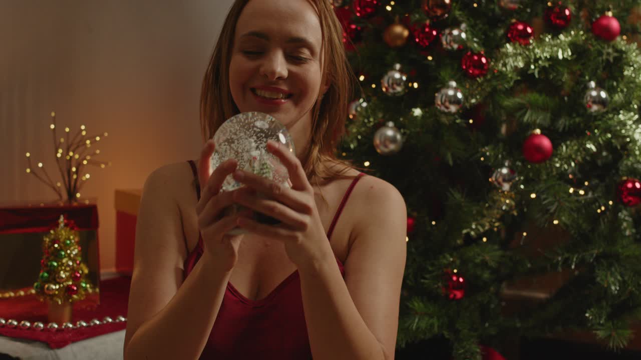A beautiful woman shakes a snow globe in slow motion and smiles in her Christmas decorated living room