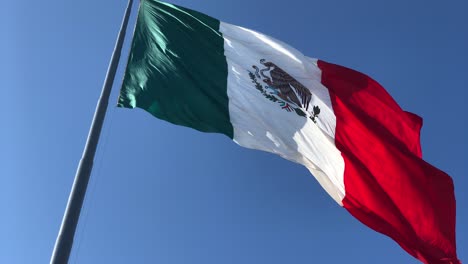 Clip-En-Cámara-Lenta-De-La-Bandera-Mexicana-Ondeando-Con-El-Viento-Mientras-Está-En-El-Poste,-Con-Un-Cielo-Azul-Claro-En-El-Fondo