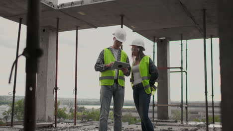 Dos-Ingenieros,-Un-Hombre-Y-Una-Mujer-Con-Cascos-Blancos-Y-Una-Tableta-En-El-Sitio-De-Construcción,-Condenan-El-Plan-De-Construcción-Del-Edificio