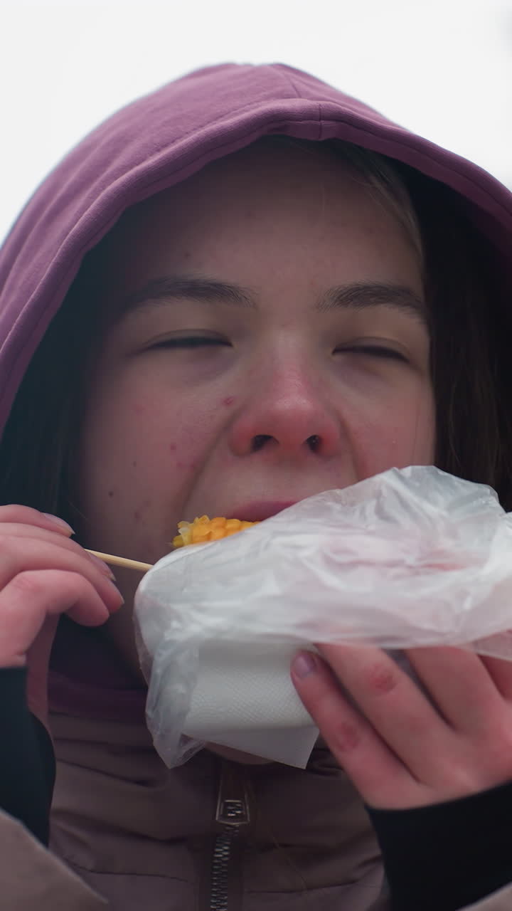 joven con chaqueta de invierno comiendo maíz, disfrutando de comida de la calle, chaqueta con capucha, bocadillo casual al aire libre, escenario de invierno, comida en palo, comiendo en clima frío, envuelto en plástico, fondo urbano