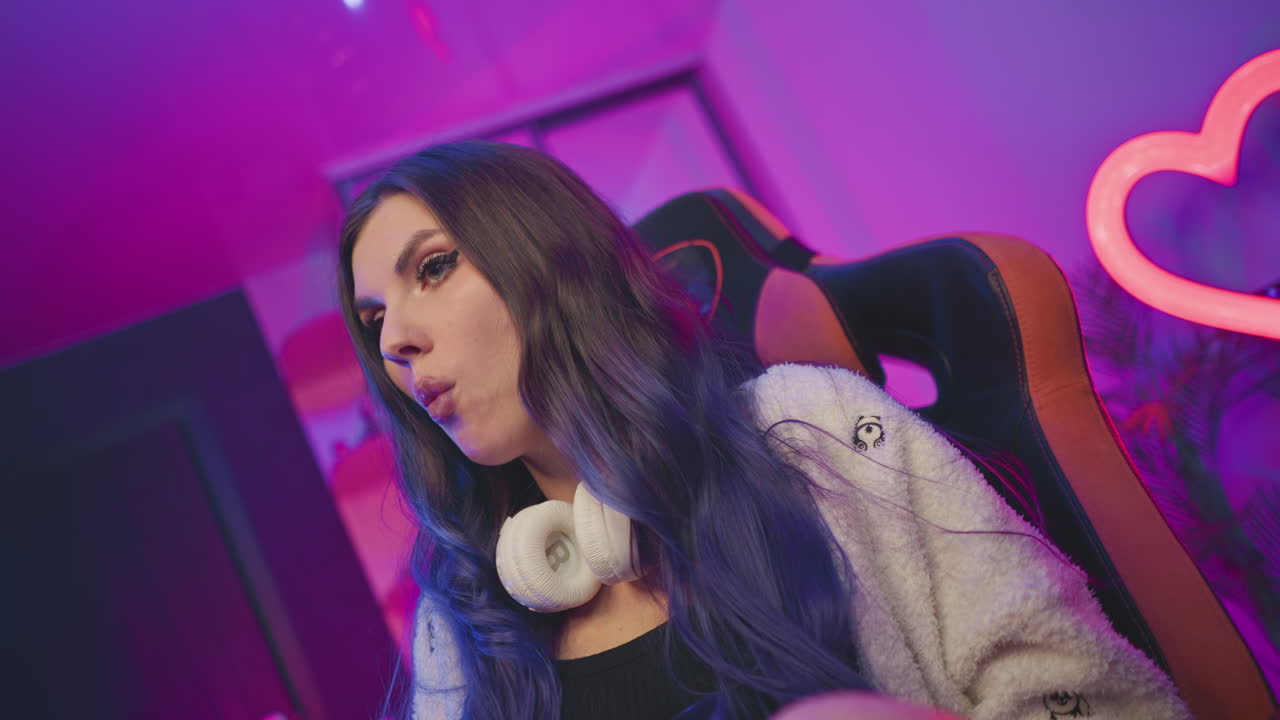 Elegant lady with long hair and headphones around neck sits on gaming chair eating popcorn with focused expression against vibrant pink and purple background lit by heart-shaped neon light