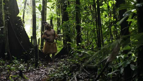 An-indigenous-guy-wearing-a-feathered-hat-and-fringed-shirt-makes-ritual-offerings-to-surrounding-trees-in-the-dense-forest-in-Leticia,-Amazon,-Colombia