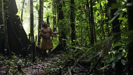 un hombre indígena con un sombrero de plumas y una camisa de flecos hace ofrendas rituales a los árboles circundantes en el denso bosque de leticia, amazonas, colombia