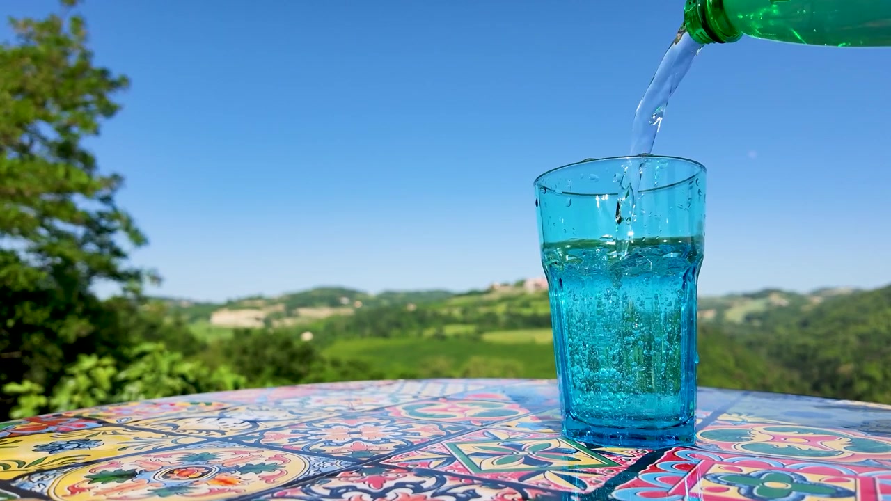 Clear sparkling water pours from a green bottle into a blue glass outdoors in sunlight
