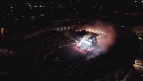 Drone-En-Las-Arenas-De-Nîmes-En-Medio-De-La-Noche,-La-Gente-Está-Viendo-El-Concierto-Y-Hay-Luces-De-Varios-Colores