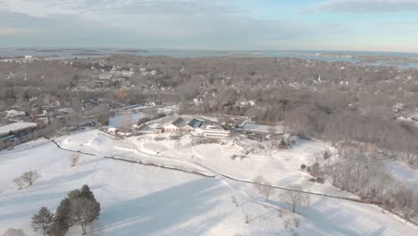 A-Una-Altura-Elevada-Y-Vista-De-Frente,-La-Vista-De-Drones-De-Un-Campo-De-Golf-Cubierto-De-Nieve-Con-La-Casa-Club-Cerca-Y-Boston-En-La-Distancia-Es-Fascinante