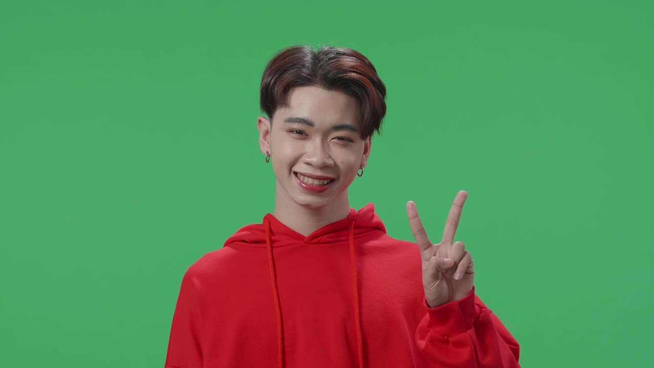 A Smiling Young Asian Transgender Male Showing Gesture Peace While Standing On Green Screen In The Studio