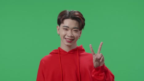 a smiling young asian transgender male showing gesture peace while standing on green screen in the studio