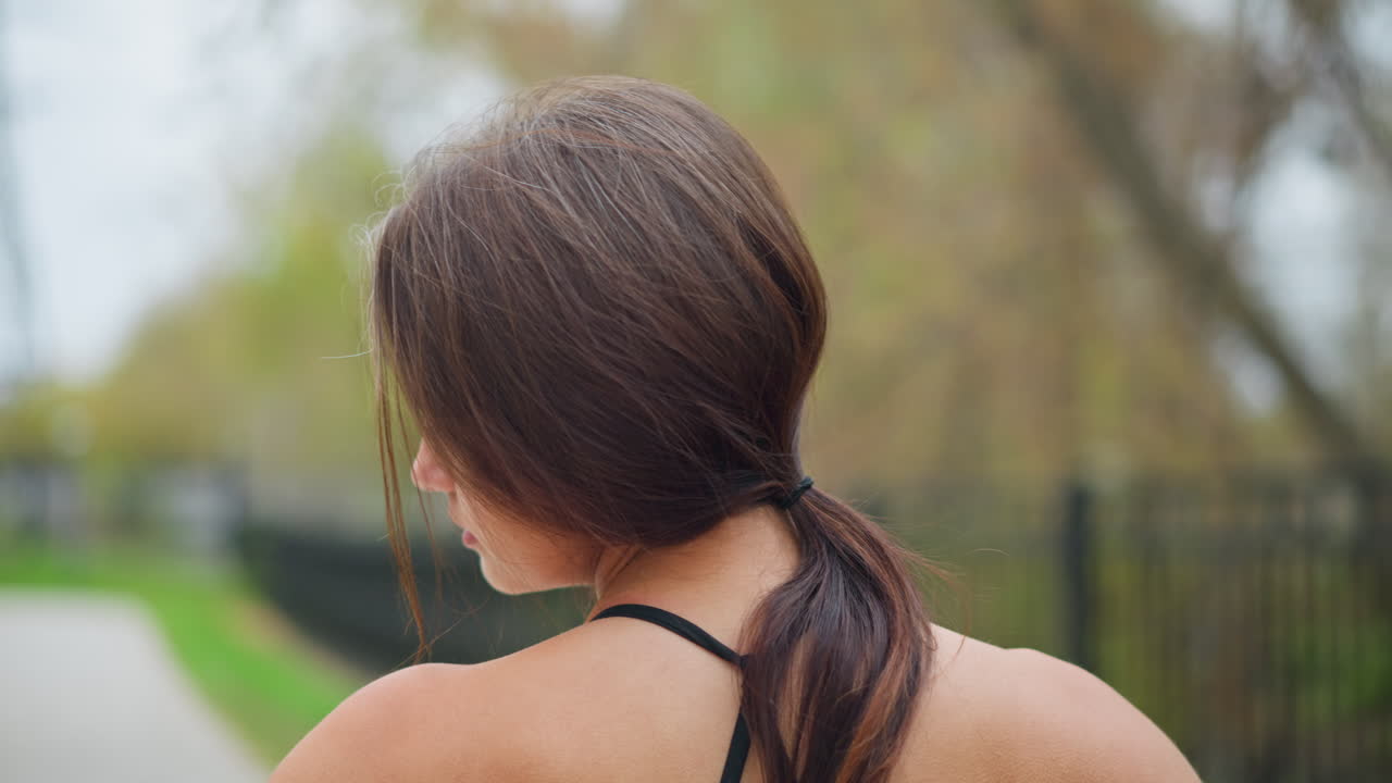 Back view of young woman performing neck stretch outdoors, making neck flexible with blurred background of trees and iron fence in park, promoting relaxation and flexibility