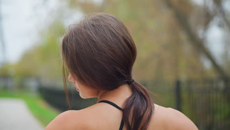 back view of young woman performing neck stretch outdoors, making neck flexible with blurred background of trees and iron fence in park, promoting relaxation and flexibility