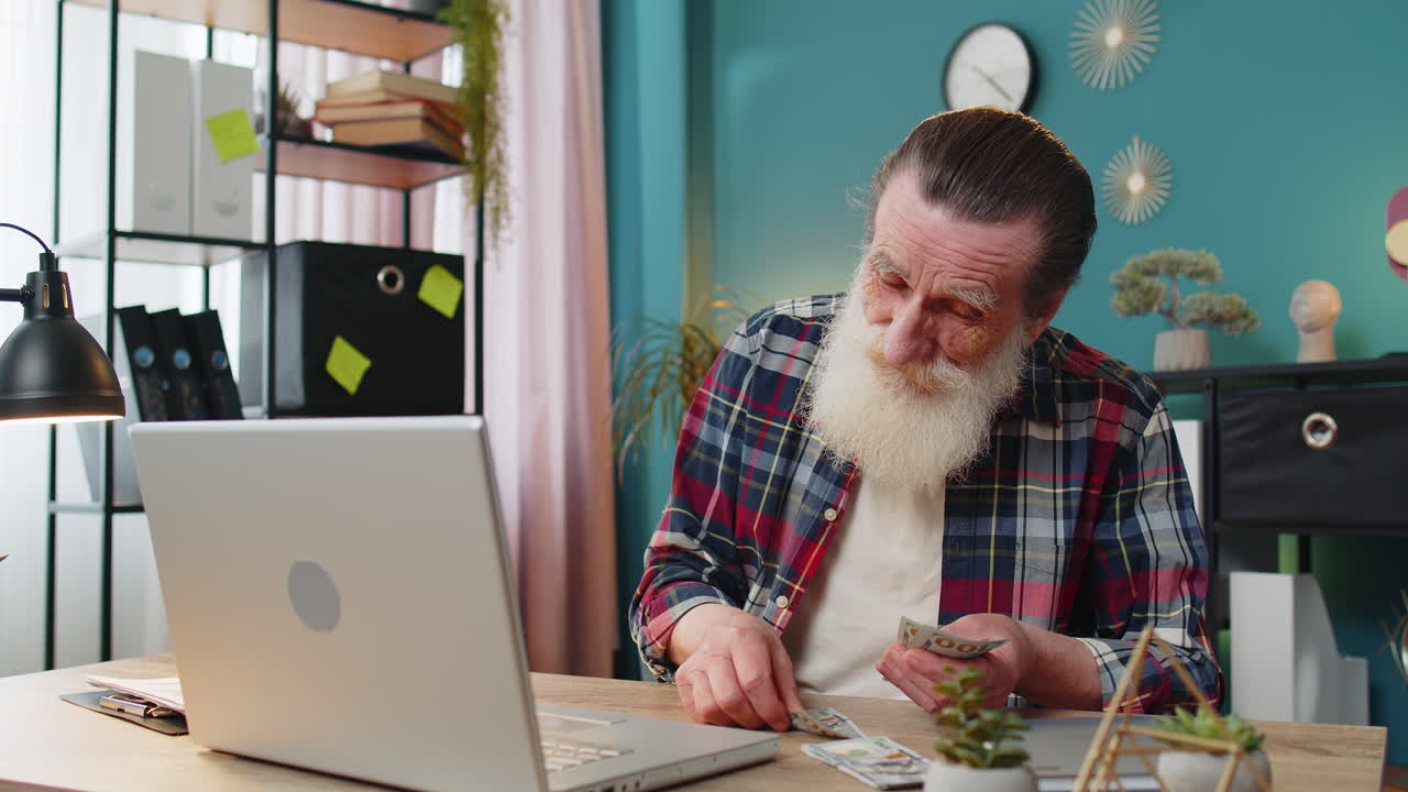 Senior man counting money cash calculates dividends profits salary and using laptop in home office