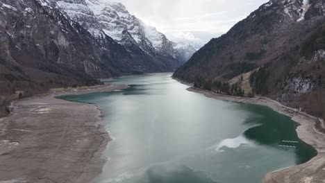 Vista-Aérea-De-Klöntalersee-Con-El-Lago-Reflejando-Las-Montañas-Nevadas-Circundantes-Y-Un-Depósito-De-Limo-Visible