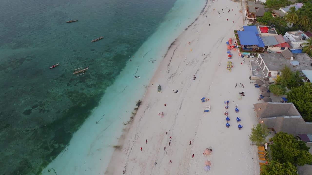 Volo cinematografico aereo con drone sopra la spiaggia di Nungwi, Zanzibar, con persone che giocano sulla costa. Acqua turchese, sabbia bianca e vivace atmosfera tropicale creano una scena di viaggio e lifestyle dinamica