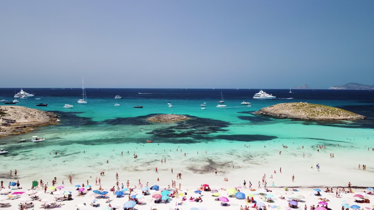 Droonikuva Formenteran saarelta Ibizan edustalta. Kamera lentää taaksepäin merelle päin kesäaikaan paljastaen rannan, ihmiset, veneet ja jahdit panoraamamaisemassa, kuvattuna keskikulmasta.