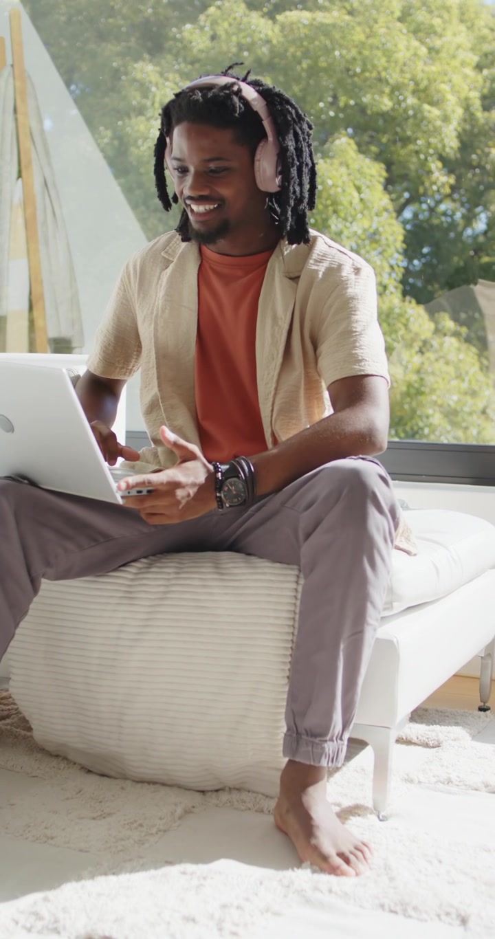 Vertical video: Man hearing audio in headphones, grinning and typing on laptop on ottoman by window