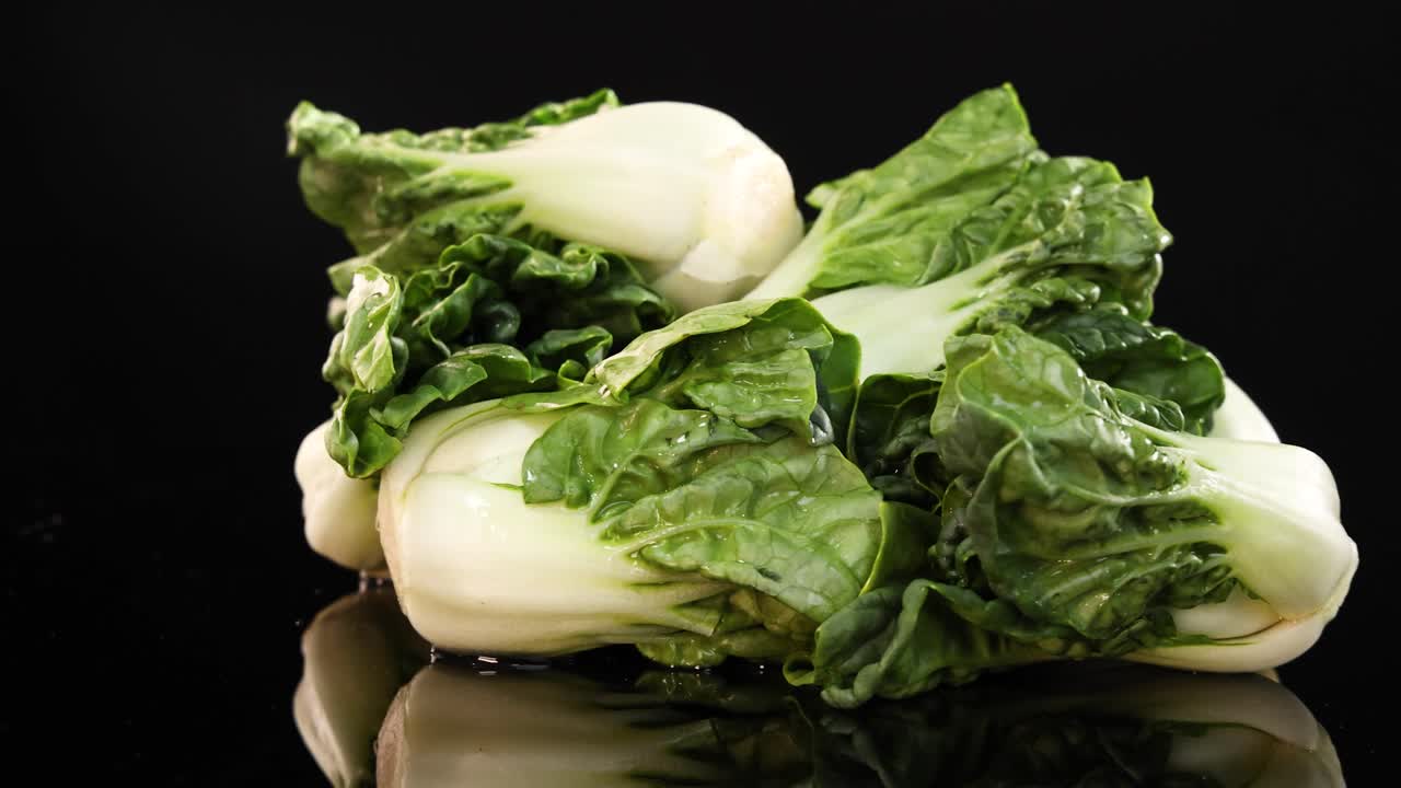 Bok choy drops onto a glossy black surface, highlighting its vibrant green leaves and crisp texture under bright lighting