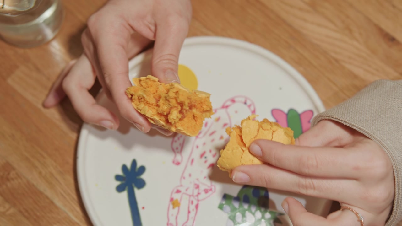 Close up of hands breaking apart yellow crumbly food item on decorative plate with colorful drawings. person wears beige sleeve and has rings interacting directly with textured snack