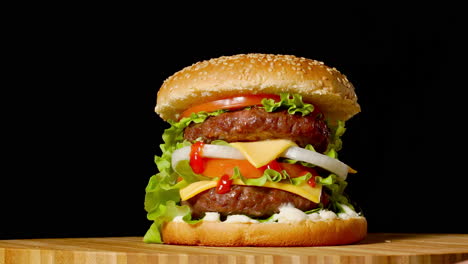 Gran-Hamburguesa-Con-Chuleta-De-Ternera,-Tomates,-Champiñones-Y-Pepinos-Con-Queso-Derretido-Gira-Sobre-Una-Tabla-De-Madera-Sobre-Fondo-Negro.