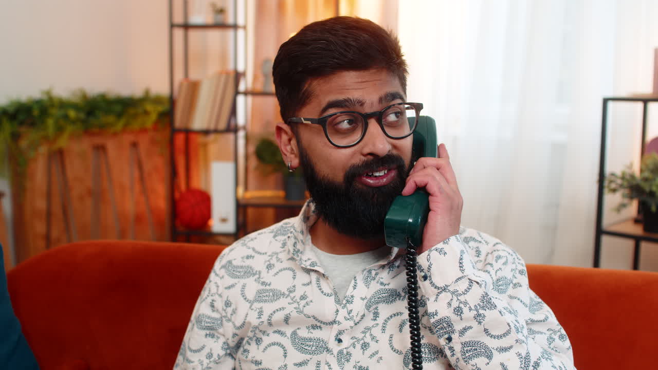 Indian man talking on retro 90s phone with excited expression receiving good news while at home sofa
