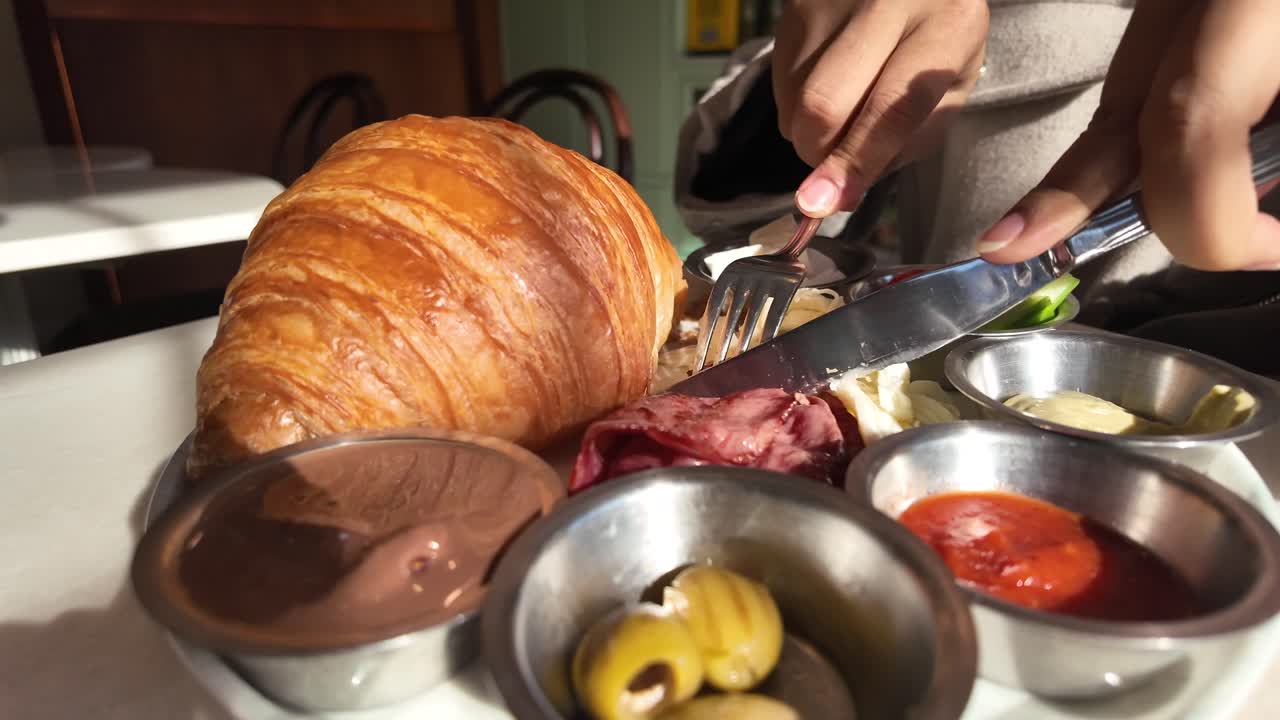Mani che tagliano carne su un piatto da colazione con croissant e salse