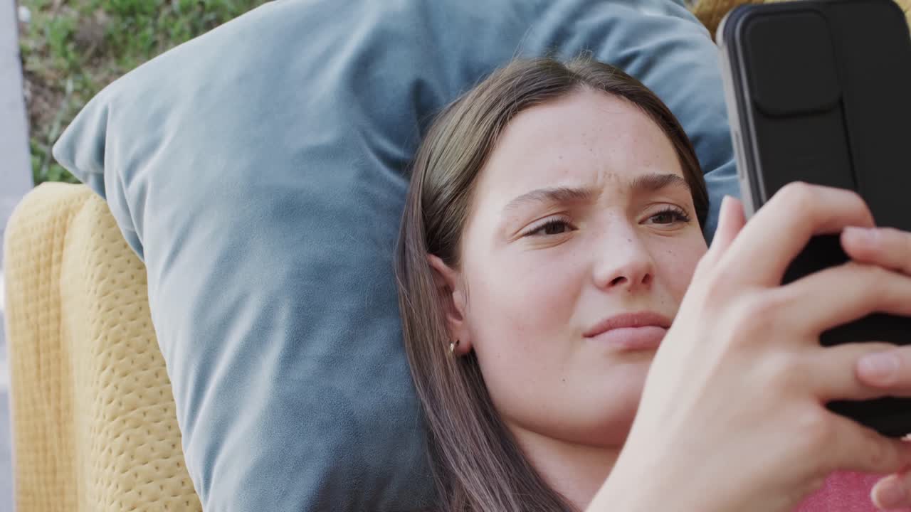 사려 깊고 화가 난 백인 십대 소녀가 스마트폰으로 문자 메시지를 보내고 슬로우 모션으로 누워 있습니다.