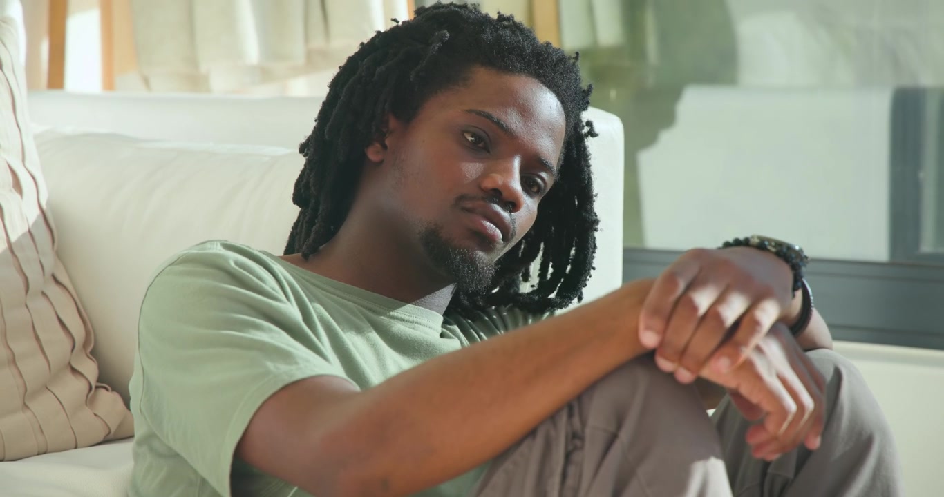 African American man on sofa at home light drawing gaze holding knees bracelet turning to window