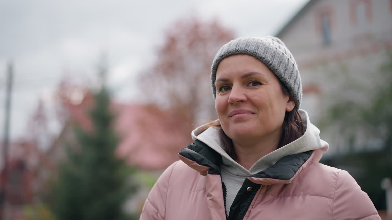 분홍색 재과 회색 베니를 입은 자신감 넘치는 여성이 장난스러운 미소를 지으며, 흐릿한 가을 잎자루, 집, 정원 배경에 대항하여 야외에 서 있습니다.