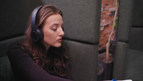 focused woman wearing headset nods gently while writing in notebook, seated in booth beside open book, tablet, and decorative tabletop plant with soft natural lighting and warm tones in background