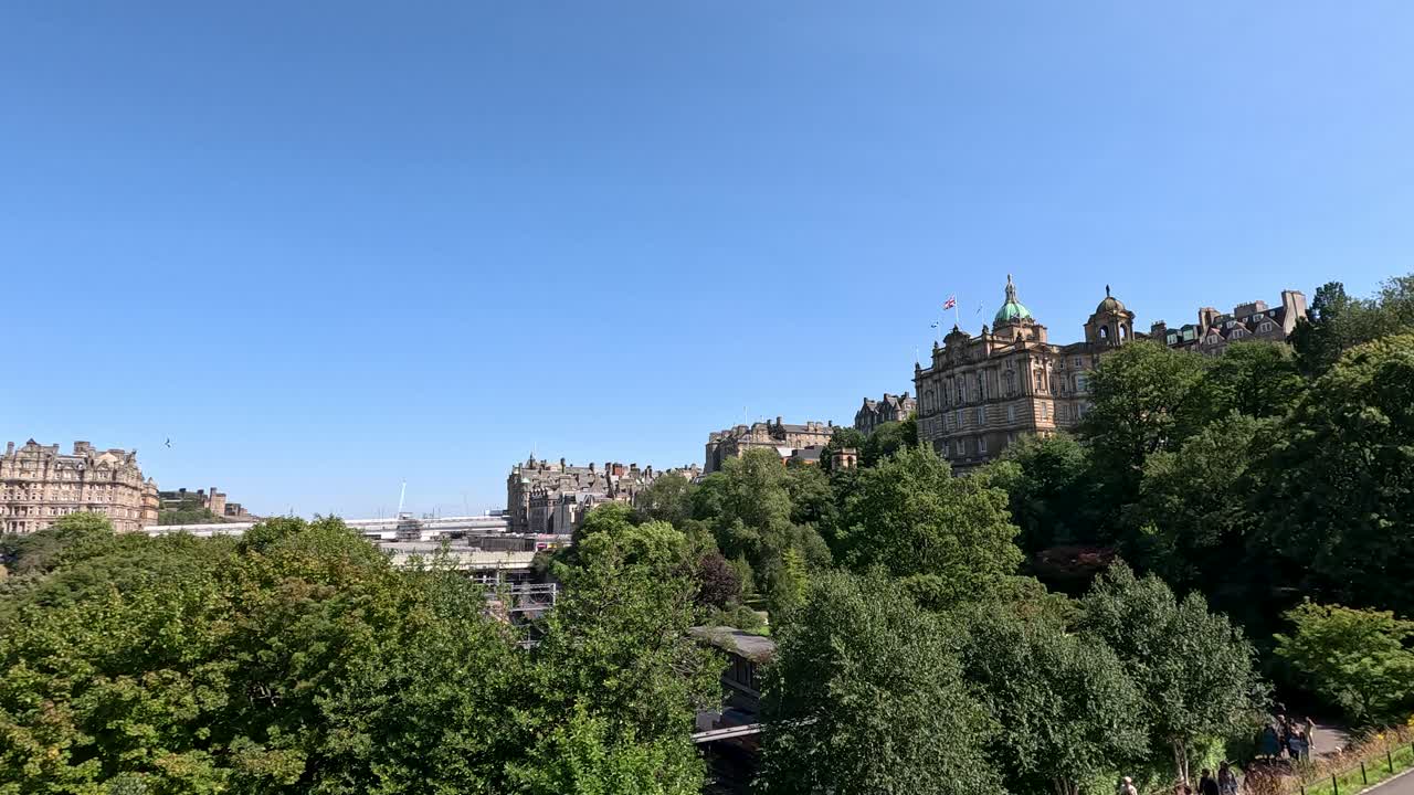 Wide daytime pan across Princes Street Gardens in Edinburgh, capturing festival crowds, historic architecture, lush greenery, and clear blue skies with smooth camera movement