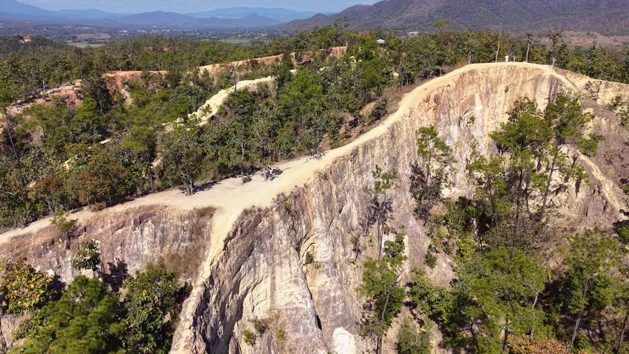 Veduta aerea con drone del Canyon di Pai nel Nord della Thailandia, che mostra drammatiche creste rosse e sentieri escursionistici tortuosi, circondato da una lussureggiante foresta, catene montuose distanti e un cielo azzurro e limpido in una luminosa giornata tropicale.