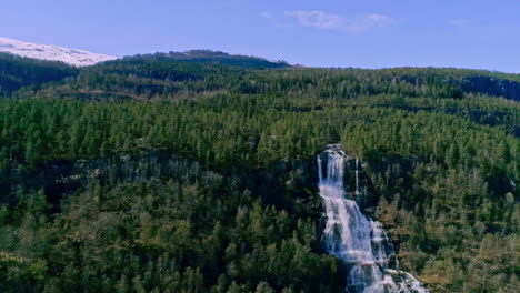 Toma-Aérea-De-Un-Dron-Hacia-Atrás-De-Una-Cascada-Desde-Un-Acantilado-De-Alta-Montaña-A-Lo-Largo-De-La-Ladera-En-Un-Día-De-Verano