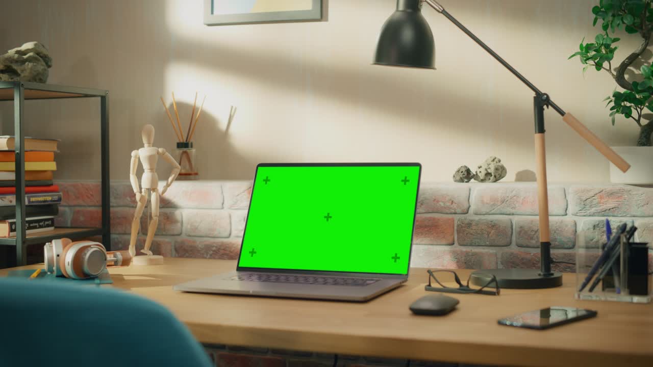 Laptop Computer Standing on a Wooden Desk with a Green Screen Chromakey Mock Up Display. Cozy Empty Loft Apartment with a Lamp, Notebooks and Smartphone on the Table. Zoom In Shot.