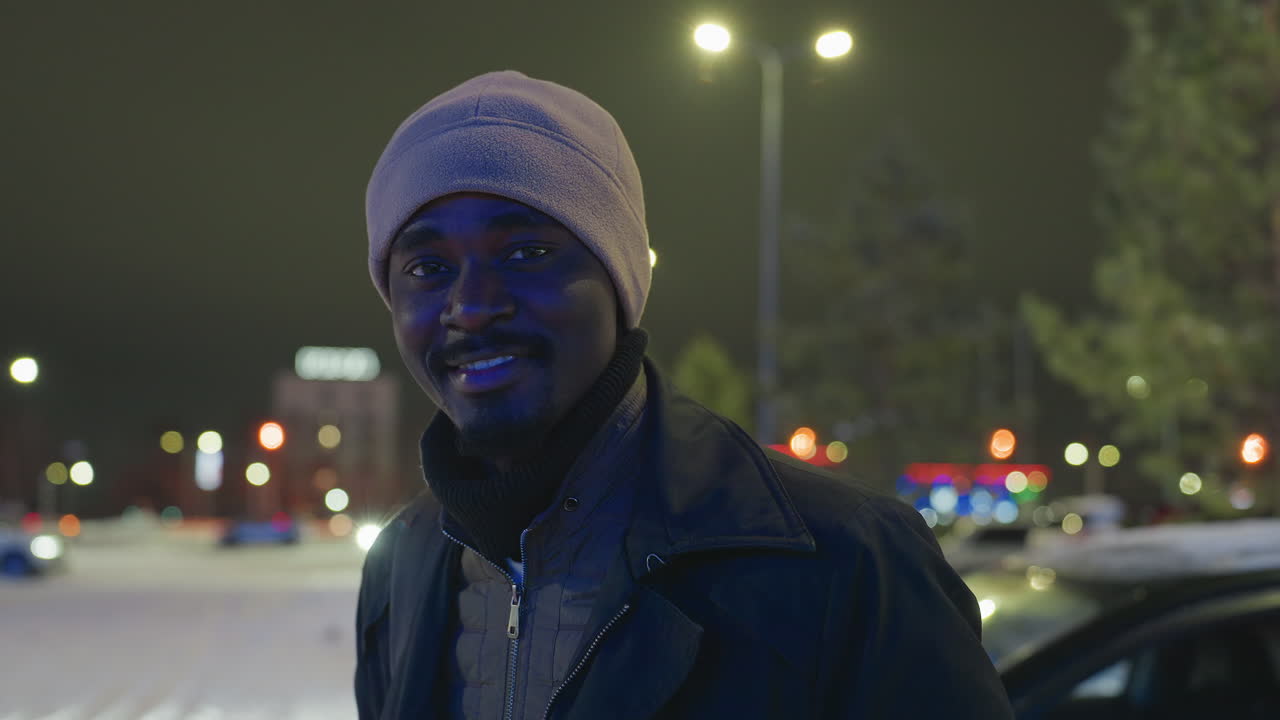 Smiling man wearing winter hat and jacket standing outdoors at night in snowy city area with blurred glowing lights, illuminated background, and relaxed expression under street lamps