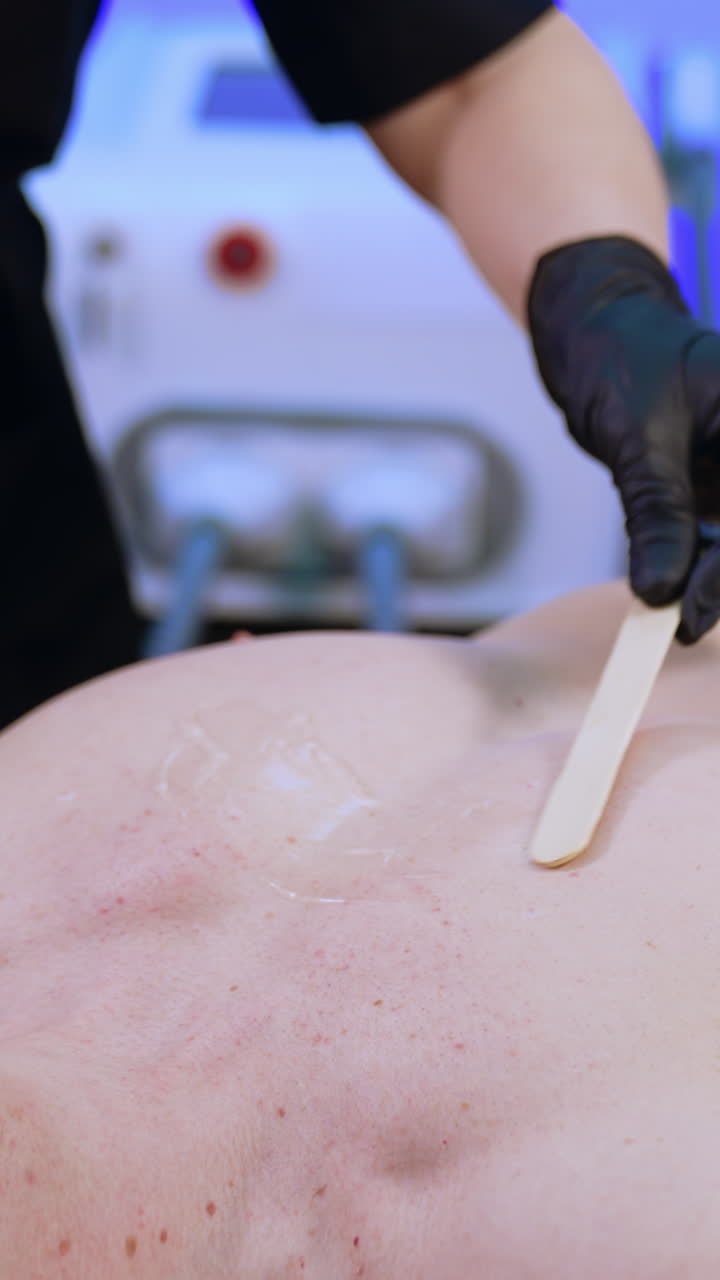 Caucasian male wearing protective goggles lies on the salon couch. Hands in black gloves apply transparent gel on man's chest. Vertical video