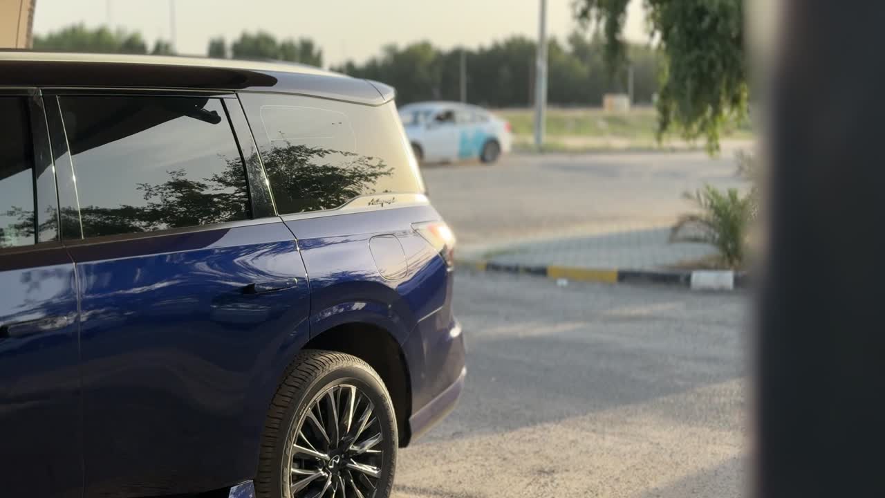 Vista posteriore laterale di un SUV blu parcheggiato all'aperto vicino a un'area stradale di giorno, con traffico sfocato, alberi e pavimentazione che creano una moderna scena di trasporto urbano.