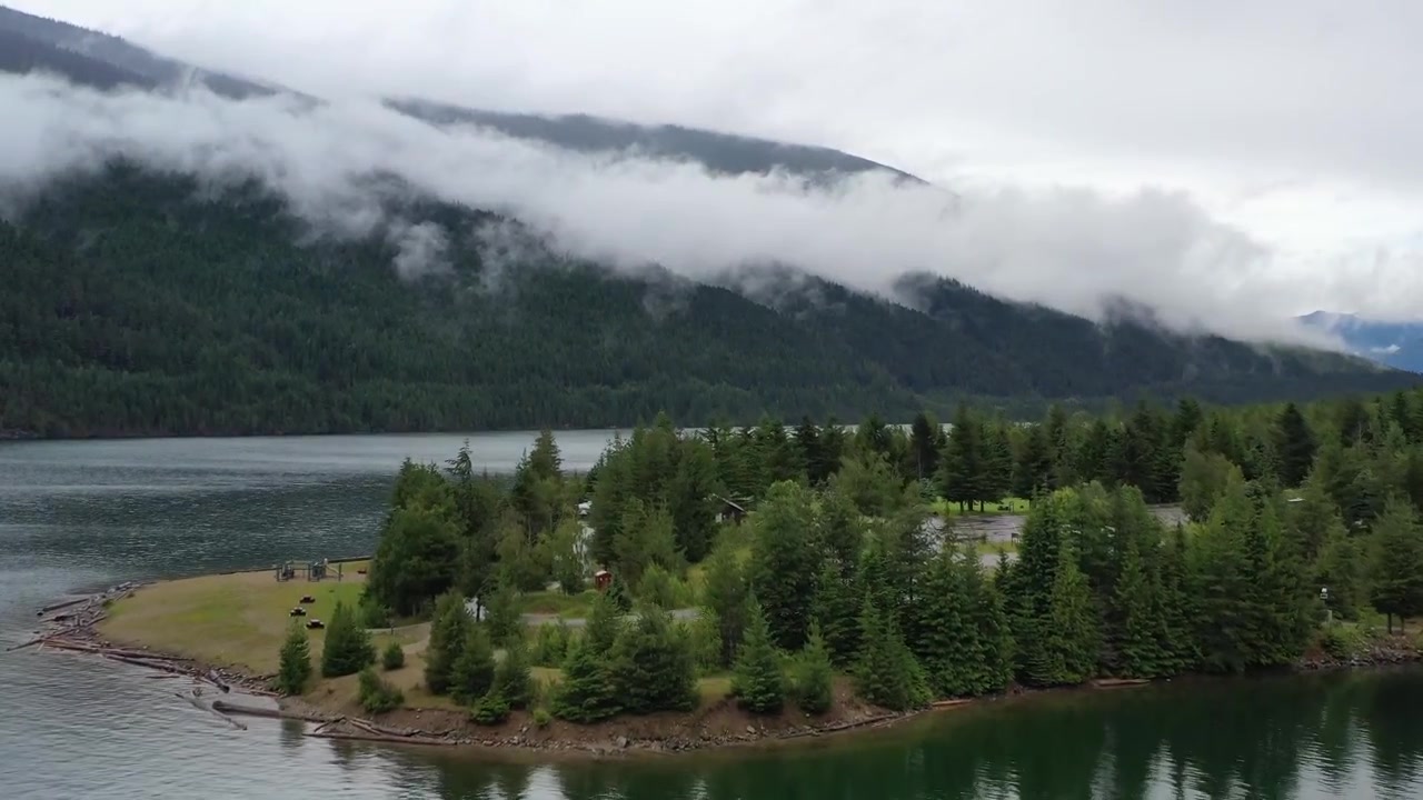 Ripresa cinematografica con drone dall'alto di una strada stretta che si snoda attraverso una fitta foresta di conifere accanto a un lago calmo e riflettente. Cime montuose suggestive e nuvole basse definiscono il paesaggio della Columbia Britannica.