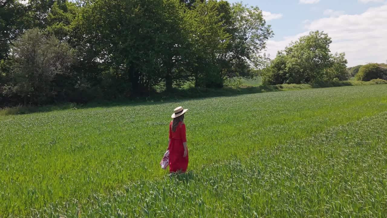 A cinematic orbit shot capturing a woman in a red dress walking gracefully through lush green meadows, surrounded by the serene beauty of the countryside