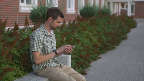 hombre sentado en un banco afuera, usando un teléfono inteligente y un reloj inteligente