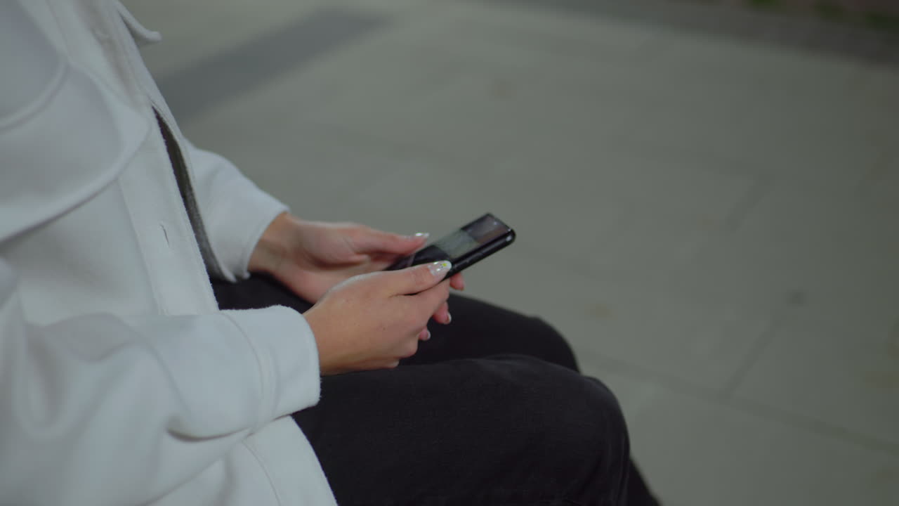 Partial view woman in white coat seated, sliding phone with neat manicure, black pants visible, background softly blurred, calm environment, focusing on device usage during serene outdoor moment at night