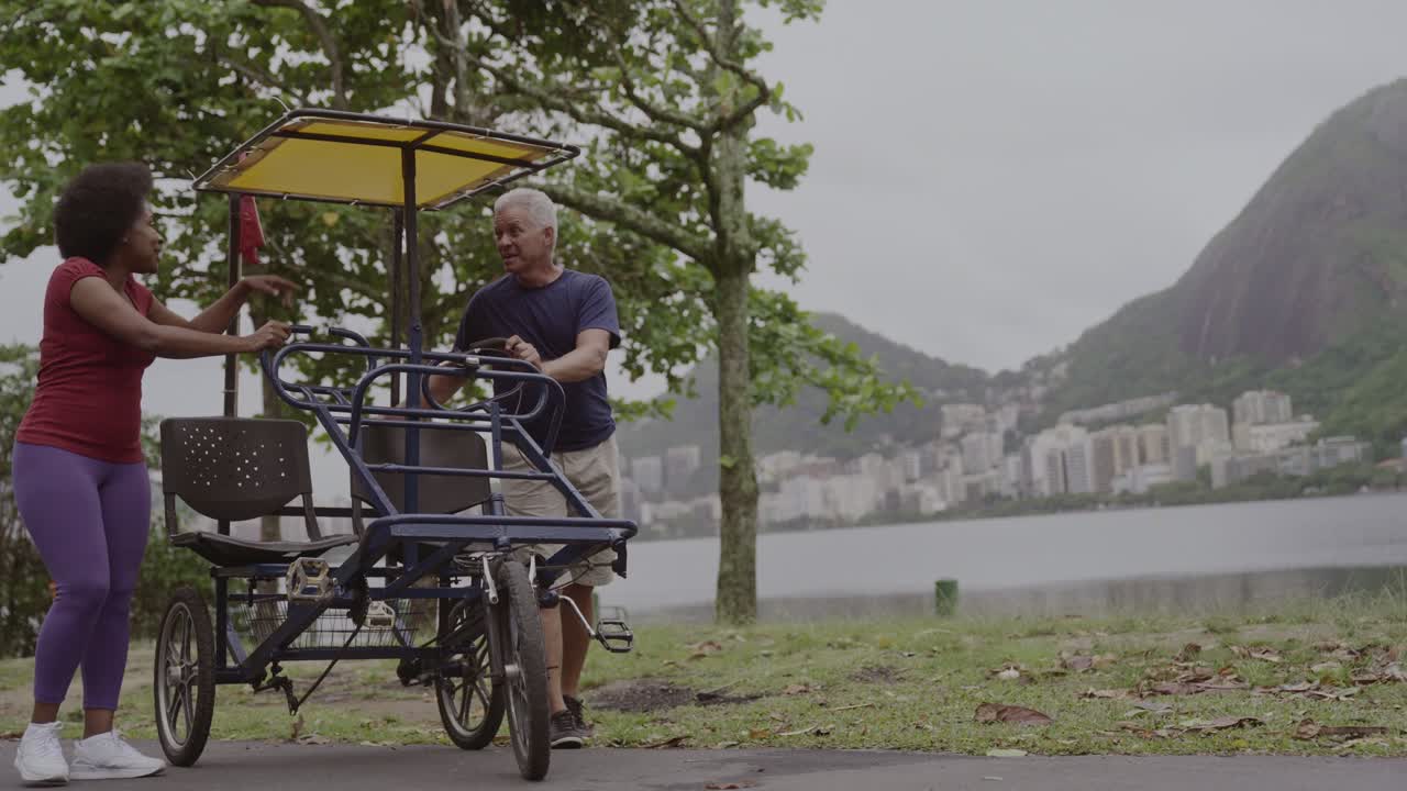 A couple with a pedal bike in a park by the lake with a city skyline and mountains in the background