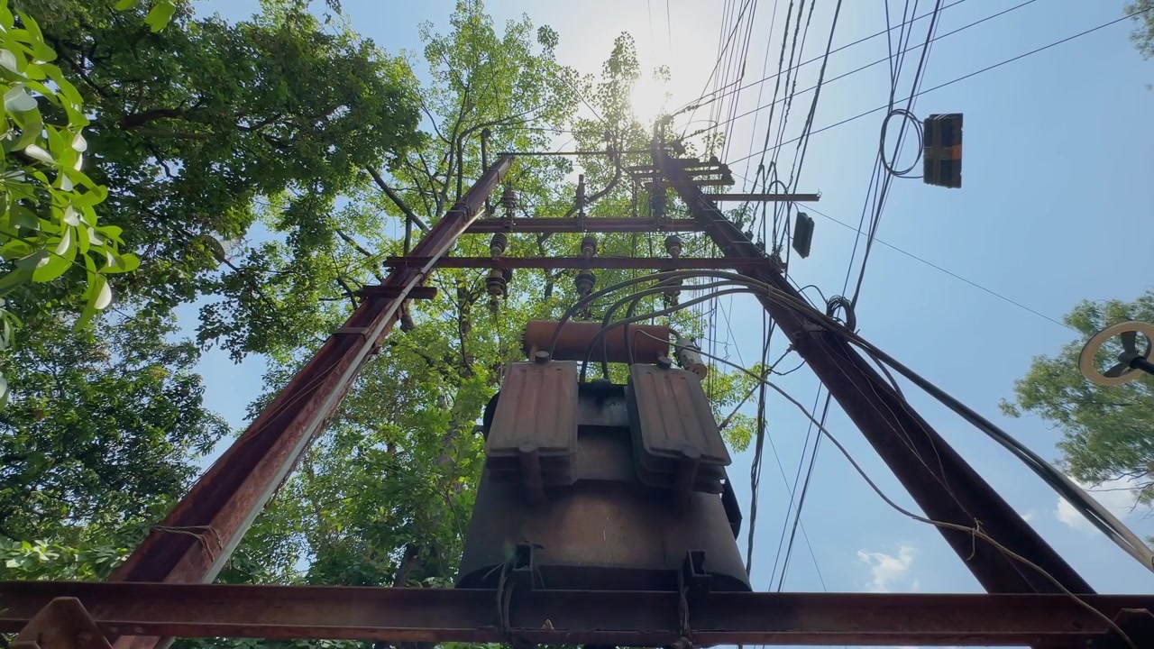 Low angle tracking shot moving beneath a pole mounted electrical transformer and overhead utility lines, highlighting power distribution infrastructure against a bright sky and tree canopy.