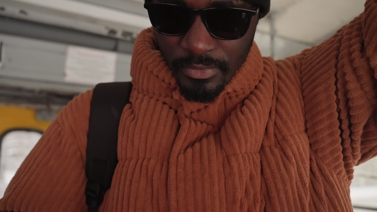Homem negro no ônibus, suéter aconchegante, gorro e óculos de sol, alça de mochila acima da cabeça, verificando a tela rachada do telefone enquanto segura o corrimão, expressão calma e confiante, luz suave da manhã, close-up de tricô texturizado.